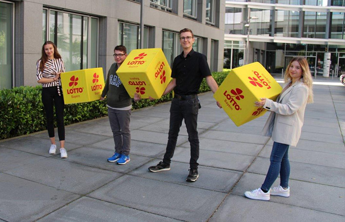 Vier Auszubildene der Staatlichen Lotterie- und Spielbankverwaltung vor der Unternehmenszentrale in München, sie tragen drei gelbe Sitzwürfel mit rotem "LOTTO Bayern"-Logo.